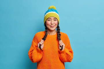 Lovely adorable ethnic girl keeps hands on pigtails being in happy mood wears knitted hat and orange sweater poses over blue background. Youth fashion emotions lifestyle concept. Fashionable teenager