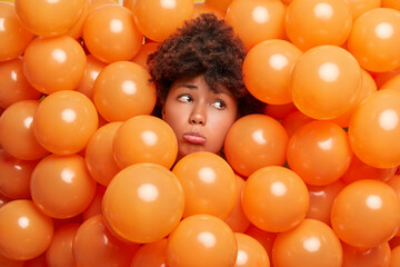 Fototapeta na wymiar Upset mournful woman with Afro hair sticks out head through inflated balloons looks sadly away doesnt want to get old surrounded by orange helium balloons being lonely on birthday celebration