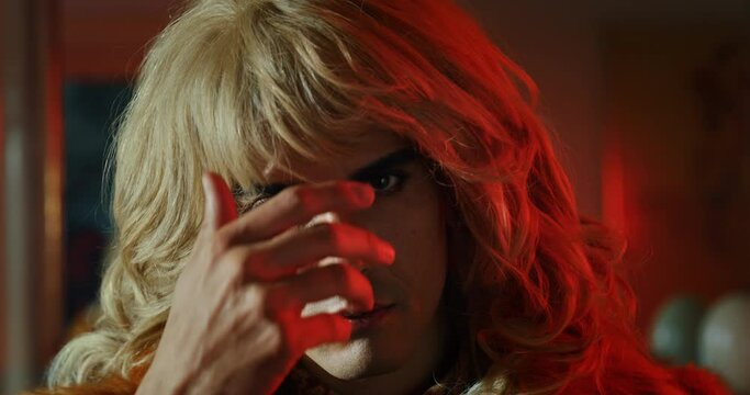 Cinematic shot of young carefree transgender woman wearing fur coat is taking off a wig and looking in camera with night club lights on background. Concept of gender expression, identity and diversity