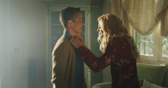 Cinematic shot of an young carefree happy smiling homosexual blond hair female gay is helping buttoning a shirt to her transgender man and giving affective kiss as sign of forever love at home.