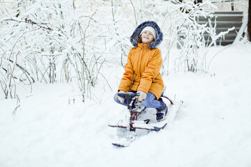 Fototapeta na wymiar Kid girl in a yellow winter park and a white knitted hat on a snow scooter with the first snow, snowfall and a walk in the park in the backyard. Frost and snowy weather.