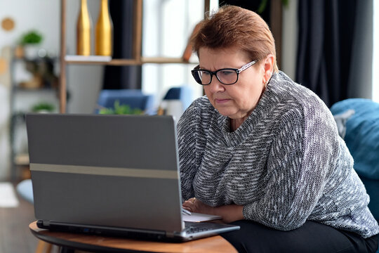 Senior Woman Using Laptop For Websurfing. The Concept Of Senior Employment, Social Security. Mature Lady Sitting At Work Typing A Notebook Computer In An Home Office.