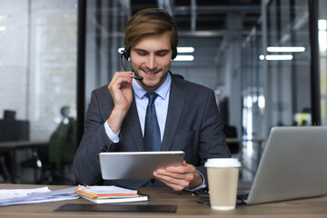 Smiling male business consultant with headphones sitting at modern office, video call looking at...