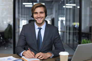 Smiling male business consultant with headphones sitting at modern office, video call looking at laptop screen. Man customer service support agent helpline talking online chat.