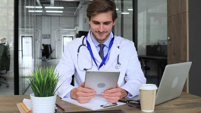Doctor in white medical uniform with statoscope reading personal file of patient on tablet and writes data to laptop.