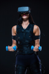 A young athletic woman in an EMS suit and virtual reality glasses with dumbbells on an isolated black background. EMS training. Electrical muscle stimulator.