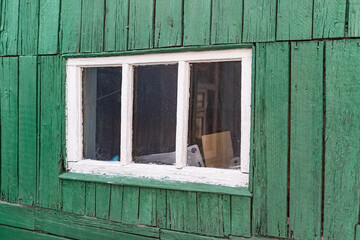 Obraz na płótnie Canvas An old wooden small window with a frame of boards painted white and thin glass. On a battered wall of wooden planks with green paint. The wall of a dilapidated building with a rough surface