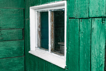 An old wooden small window with a frame of boards painted white and thin glass. On a battered wall of wooden planks with green paint. The wall of a dilapidated building with a rough surface