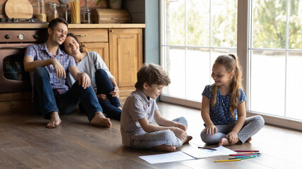 Wide banner view of happy young Caucasian family with two little kids sit on warm floor in new home kitchen relaxing. Smiling parents enjoy weekend indoors rest with small children painting drawing.