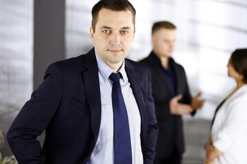 Fototapeta premium Portrait of a self-confident middle aged businessman in a blue suit, standing in a modern office with his colleagues at the background. Concept of business success