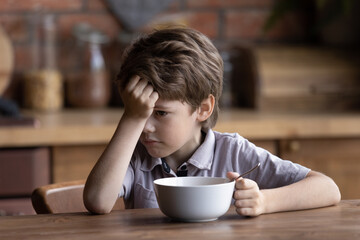 Fototapeta na wymiar Unhappy small Caucasian boy child sit at kitchen table have no appetite for healthy breakfast. Sad upset little 7s kid refuse to eat nutritious meal or food at home. Children healthcare concept.
