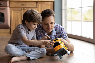 Loving young Caucasian father lying on floor feel playful teach little son repair toy car. Happy caring dad and small boy child have fun fixing automobile at home together, play enjoy family weekend.