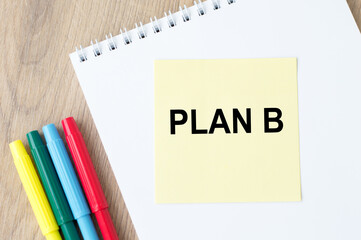 plan B on yellow note paper, which lies on a notebook on the table, next to the colored markers.