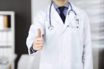 Unknown male doctor standing with thumbs up sign in clinic near his working place, closeup. Perfect medical service in hospital. Medicine and healthcare concept