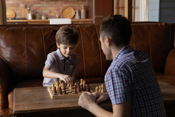 Small smart Caucasian boy child have fun playing chess with caring young dad at home. Focused little kid engaged in board game with father, think ponder over strategy. Children development concept.