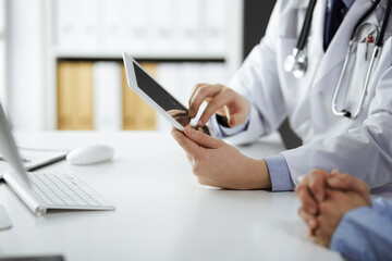 Unknown male doctor and patient woman discussing current health examination while sitting in clinic and using tablet computer, closeup of hands. Medicine and healthcare concept