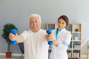 Experienced female doctor supports senior man's arms as he lifts dumbbells in medical office.