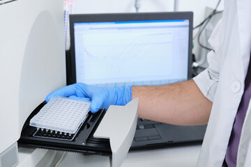 A researcher putting PCR plate on the thermal cycler for DNA amplification. Curve chart on monitor behind. Coronavirus PCR test.