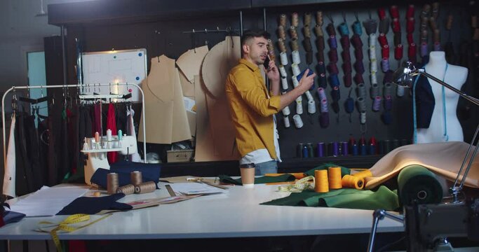 Caucasian young happy man fashion designer chatting on smartphone with client in workshop and choosing sewing thread. Cheerful male professional speaking on cellphone. Fashion industry concept