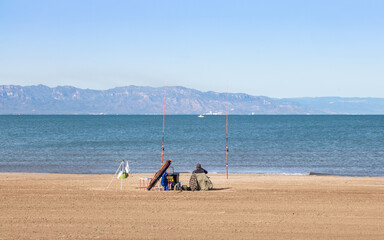 Man fishing at the beach with two rods and other fishing gear, land based fishing is a relaxing hobby. Mountains and boats at the background. Delta del Ebre, Mediterranean, Tarragona, Catalonia, Spain