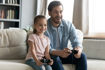 Smiling young father sitting on sofa with happy small 7 years old daughter, holding controllers in hands involved in funny childish video game in living room., family weekend leisure pastime concept.