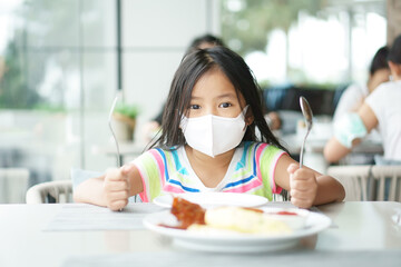 Asian child or kid girl wearing white face mask to holding spoon and fork wait eat delicious food...