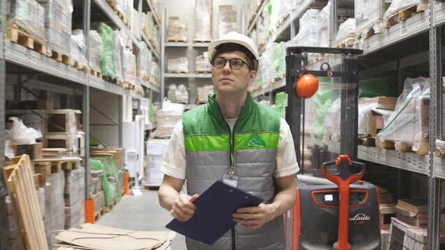 Caucasian warehouse worker is checking his list on clipboard in warehouse, transportation logistics by trucks. Wholesale, logistics, export concept.