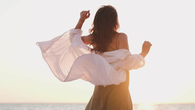 beautiful girl dancing on the beach in a white shirt. Free girl with long hair in a white shirt dances in the sunset. Healthy woman dancing on the beach.