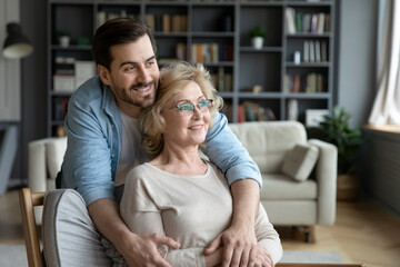 Smiling young man cuddling calm peaceful older mature mother seated on chair in living room. Happy different generations family enjoying dreamy mindful moment, thinking of future at home, copy space.