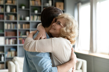 Side view affectionate young man embracing happy middle aged 60s mother, visiting at home. Loving...