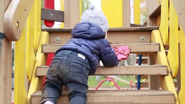 Funny little boy in a warm jacket and hat crawls, climbs and climbs up a wooden ladder on the Playground in the Park. The kid got all dirty playing in the street. The concept of a happy childhood