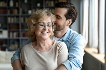 Happy middle aged older woman in eyeglasses standing with closed eyes, enjoying tender loving moment with grown son at home. Devoted young man showing care to smiling retired old mother indoors.