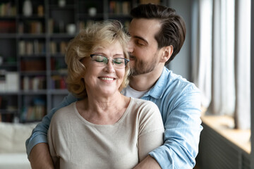 Fototapeta premium Happy middle aged older woman in eyeglasses standing with closed eyes, enjoying tender loving moment with grown son at home. Devoted young man showing care to smiling retired old mother indoors.