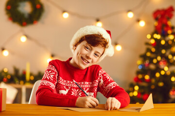 Smiling boy in red hat and sweater sitting and writing letter to Santa with wishes and dreams