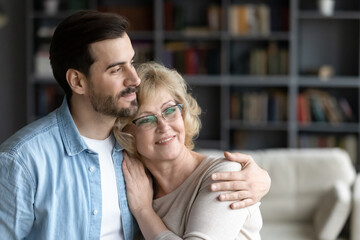 Dreamy young bearded man cuddling happy older 60s retired mother, visualizing future together. Thoughtful smiling different generations family recollecting memories, enjoying calm moment at home.