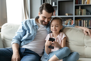 Obraz premium Little cute baby girl showing funny mobile application to affectionate caring young father, relaxing together on sofa at home. Happy young dad watching small daughter playing game on smartphone.