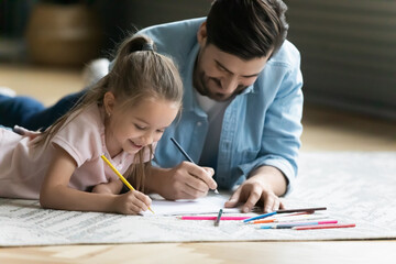 Happy small 7 years old adorable kid girl involved in pictures drawing in paper album with caring young father, lying together on floor carpet in living room, family weekend creative activity pastime.
