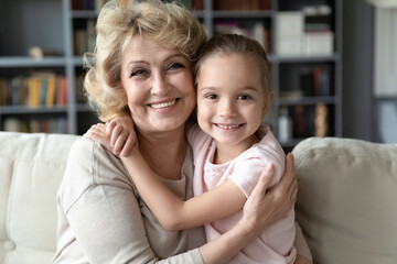 Portrait of happy middle aged older woman embracing small adorable 7 years old granddaughter, relaxing together on comfortable sofa at home. Bonding devoted multigenerational family looking at camera.