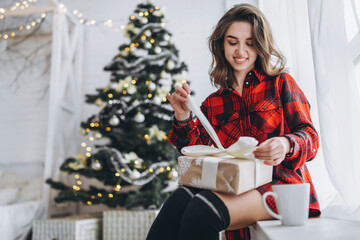 Pretty woman in shirt and socks sitting on the windowsill with christmas gift box, new year tree behind.