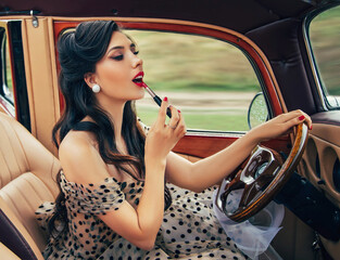 Distracted driver. Young attractive retro woman looking in rear view mirror painting her red lips...