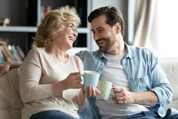 Joyful different generations bonding family relaxing on cozy sofa, drinking hot tea coffee, involved in funny conversation. Happy older 60s retired mother talking having fun with grown son at home.