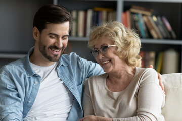 Head shot happy young bearded man embracing beautiful smiling middle aged senior mother in eyewear, relaxing together on comfortable sofa in living room, enjoying funny conversation or gossiping.