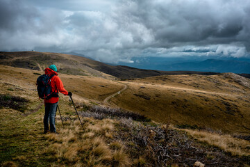 A hiking man in a red jacket standing below the dramatic grayish sky and watching autumn-colored mountain peaks and fields