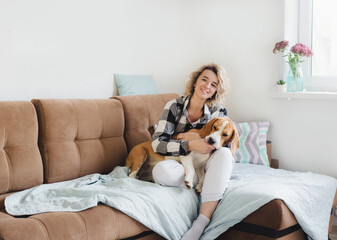 Happy young woman putting down a Beagle dog while sitting on a sofa in a checked shirt. The youth way of life, the concept of home and animals.