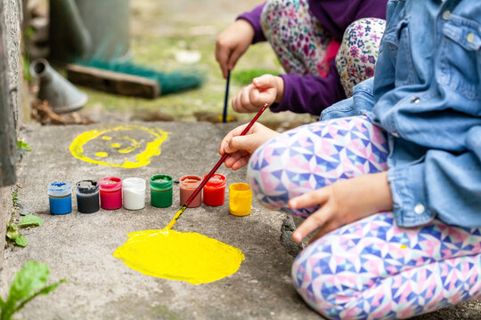 Young children painting on concrete, two anonymous girls engaging in art therapy activities. Small kids spreading colorful paint on the ground using a brush, outdoors scene, closeup. Kids and art