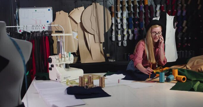 Beautiful young happy Caucasian woman fashion designer chatting on mobile phone with client in her own small workshop. Cheerful female dressmaker speaking on cellphone and taking sewing thread