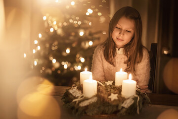 Child sits alone in front of a glowing Advent wreath and looks forward to Christmas. Girls,...