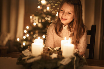 Child sits alone in front of a glowing Advent wreath and looks forward to Christmas. Girls,...