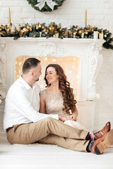 Obraz premium Merry Christmas. Young couple celebrating Christmas at home near fireplace. Happy young couple sitting at home near christmas tree. Picture of young couple hugging in Christmas time