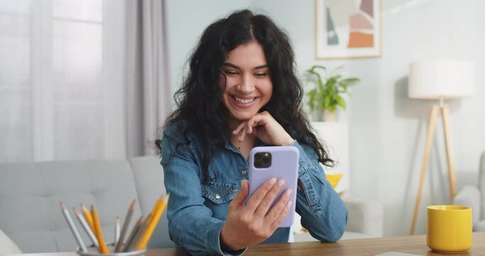 Pretty Caucasian woman sitting at table and video calling friend in home office. Attractive young female talking on smartphone facetime app and laughing indoors. Technology, social media concept.